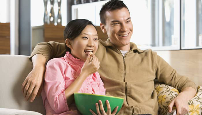 Married Asian couple watching tv on the couch and eating popcorn