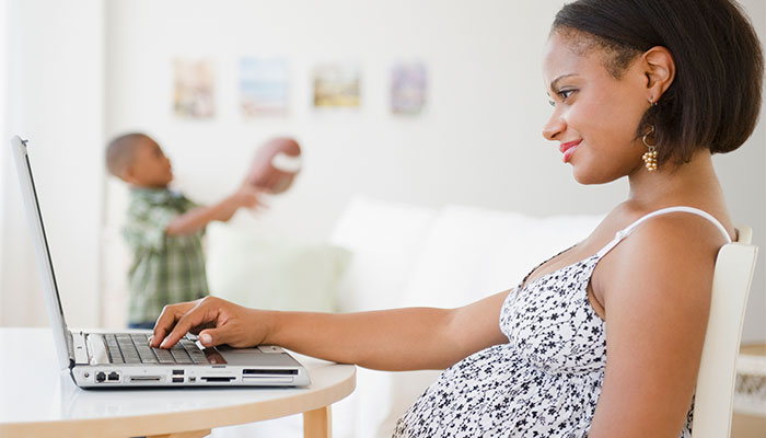 Pregnant woman uses laptop while child plays in background